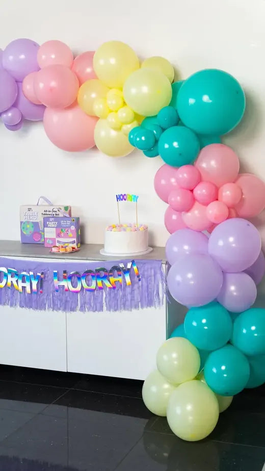 A pastel balloon arch around a party table with a Hooray banner